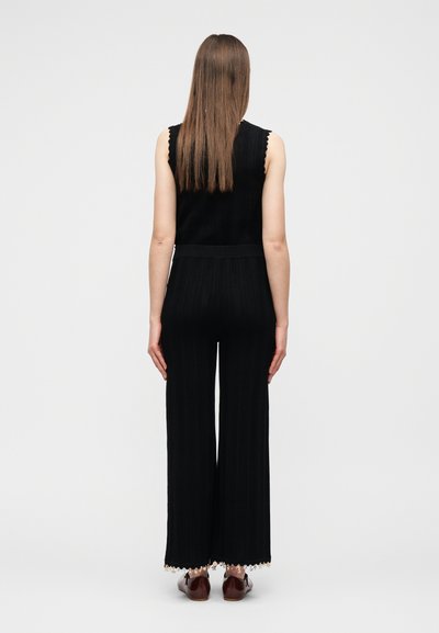 Γυναίκα που στέκεται με την πλάτη γυρισμένη, φοράει μαύρο αμάνικο τοπ και παντελόνι φαρδύ, με καφέ loafers και ίσια καστανά μαλλιά.