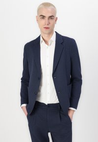 Blazer texturizado azul marino con dos botones, solapas con muesca y bolsillos con solapa, llevado sobre una camisa blanca de botones. Los pantalones también son azul marino.