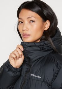 Moteris su tamsiais plaukais užsisega juodą "Columbia" pūkinę striukę su aukštu apykakle priešais šviesų foną.