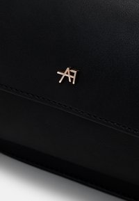 Petit logo métallique doré "A7" sur une surface lisse en cuir noir avec une couture fine visible sous le logo.