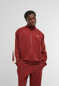 Muž v bordovom športovom obleku s bielymi pruhmi na rukávoch a zodpovedajúcich nohaviciach, stojaci s rukami v vreckách na bielom pozadí.