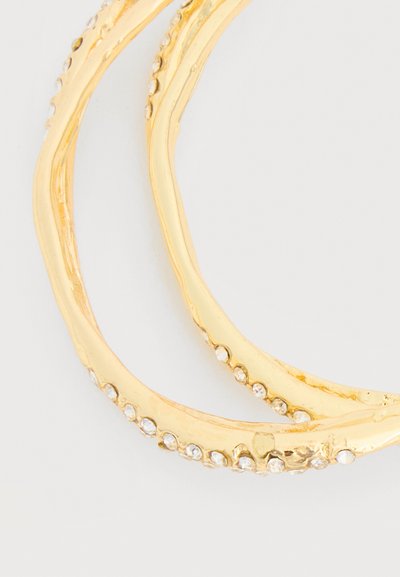 Zlati obroči za ušesa z majhnimi prozornimi kristalčki, vgrajenimi vzdolž zunanje robove, postavljeni na svetlo ozadje.