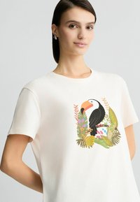 Balta medvilninė marškinėliai, kuriuose pavaizduota spalvinga tukano grafika, apsupta tropinių lapų ir gėlių su žaliais, rožiniais ir oranžiniais akcentais.