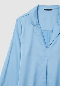 Blusa azzurra chiara realizzata in tessuto liscio. Presenta un colletto, maniche lunghe e una piega frontale con dettagli minimi. Etichetta visibile all'interno.