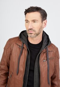 Ruskea nahkatakki, jossa on harmaa huppulining, vetoketjulliset taskut ja ribattu hihansuu. Päällä musta crewneck-paita.