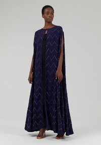 Langer marineblauer Umhang aus Stoff mit Zickzackmuster, mit metallischen Akzenten. Seitliche Schlitze und ein minimalistisches Kragen-Detail werten das Design auf.