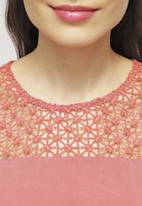 Koralfarvet kjole med en tekstureret blonde yoke, der har geometriske mønstre og et glat, ensfarvet stof forneden. Rund halsudskæring, korte ærmer.