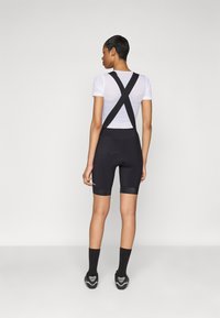 Black cycling shorts with suspenders, smooth texture, paired with a white mesh top. Notable X-back design. Cycling shoes and black socks.