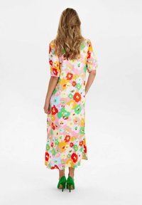 Kvinna med långt vågigt hår som bär en färgglad blommig midiklänning och gröna klackar, stående med en hand på höften och vänd bort.