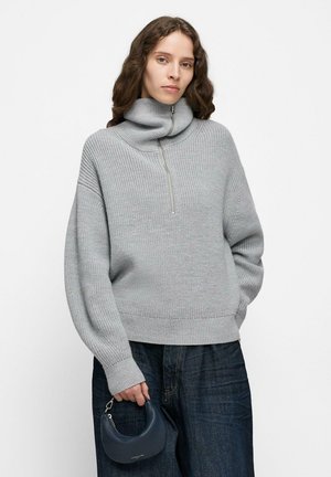 Frau mit welligem Haar, die einen grauen Rollkragenpullover mit Reißverschluss und dunkle Jeans trägt, in einer Hand eine kleine marineblaue Halbmond-Handtasche haltend.