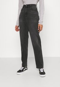 Jeans de mezclilla negros de cintura alta con corte recto, que cuentan con bolsillos frontales y cierre de botón, combinados con zapatillas negras y blancas.