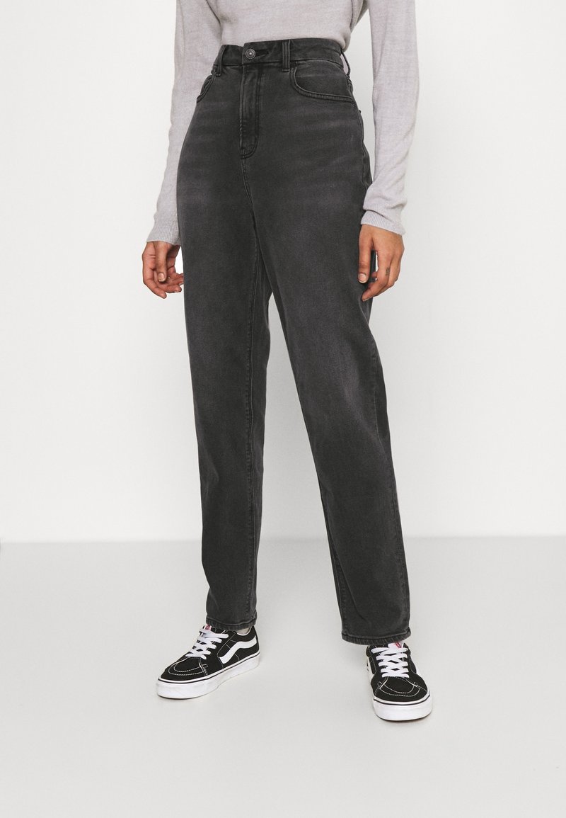 Jeans de mezclilla negros de cintura alta con corte recto, que cuentan con bolsillos frontales y cierre de botón, combinados con zapatillas negras y blancas.