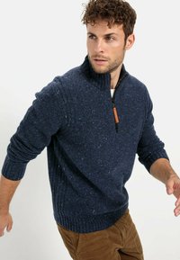 Granatowy sweter ze splotem z wysokim kołnierzem, z półsuwnym zapięciem, ściągaczami na rękawach i teksturowanym wykończeniem, noszony na beżowych spodniach.