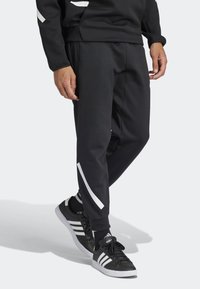 Person som bär svarta smalnande sweatpants med vit diagonal rand, svarta Adidas-sneakers med vita ränder, och en svart sweatshirt.