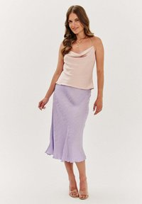 Světlé růžové saténové cami s hlubokým výstřihem, spárované s lila midi sukní s texturou a rozšířeným lemem. Průhledné páskové lodičky.