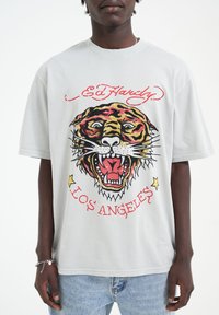 Šedé bavlněné tričko s velkým barevným grafickým návrhem tygra a červeným textem "Ed Hardy" nahoře. Má krátké rukávy a volný střih.