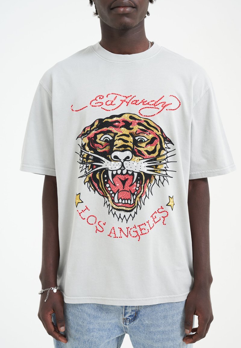 Šedé bavlněné tričko s velkým barevným grafickým návrhem tygra a červeným textem "Ed Hardy" nahoře. Má krátké rukávy a volný střih.