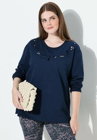 Top de manga larga azul marino con detalle bordado en el cuello, combinado con leggins estampados. La modelo sostiene un bolso texturizado en color beige.