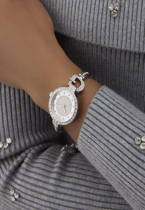 Silberne Armbanduhr mit rundem Zifferblatt, verziert mit Strasssteinen. Verfügt über ein silbernes Armband und ein Schließdesign, getragen auf einem grauen Rippensweater.