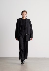 Melns uzvalks ar strukturētu žaketi ar sudraba pogām un platām biksēm, kombinēts ar teksturētu melnu topu un šaurām melnām kurpēm.