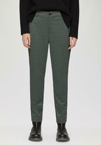 Pantalons à motifs verts présentant un design géométrique, coupe slim, poches et fermeture à bouton, associés à des bottines noires.