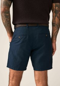 Marineblaue Shorts aus strukturiertem Stoff; mit zwei Gesäßtaschen mit Knöpfen und einem gewebten braunen Gürtel. Sichtbare Tattoos am Arm.