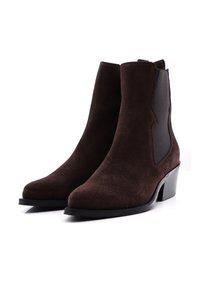 Un par de botas de ante marrón oscuro hasta el tobillo con paneles elásticos laterales negros y tacones bajos en un fondo blanco.