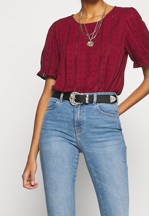 Bordeauxrood blouse met korte mouwen en textuurrijke details, gecombineerd met lichtblauwe, hooggeknoopte spijkerbroek en een zwarte riem met een zilveren, sierlijke gesp.