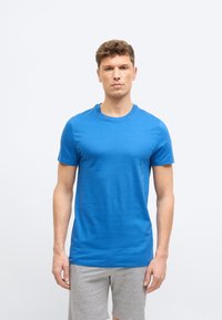 Νεαρός άνδρας που στέκεται, φορώντας στενή μπλε κοντομάνικη μπλούζα και ανοιχτόγριες σορτς, μπροστά σε απλό λευκό φόντο.