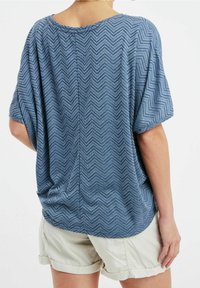 Top azul suave de manga corta con patrón de espiga, corte relajado y cuello redondeado; cuenta con un detalle de costura en la espalda y un dobladillo ligeramente curvado.