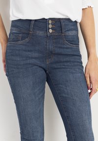 Höga blå denimjeans med en slät yta, prydda med tre metallknappar och kontrasterande sömmar; inkluderar sidofickor.