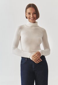 Maglione bianco a collo alto con maniche lunghe e colletto a coste, abbinato a jeans blu scuro. Tessuto morbido, vestibilità slim e design senza cuciture.