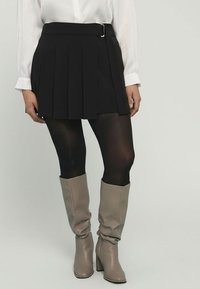 Schwarzer, plissierter Mini-Rock mit einem Gürtel und silberner Schnalle, kombiniert mit schwarzen Strumpfhosen und beigen knielangen Stiefeln mit einem leichten Glanz.