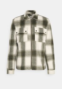 Chaqueta de cuadros con una mezcla de colores verde y blanco, elaborada en una tela suave. Cuenta con cierre de cremallera y dos bolsillos en el pecho con botones a presión.