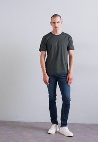 Homme adossé à un mur gris clair uni, portant un t-shirt gris foncé, un jean bleu et des baskets blanches, les mains détendues le long du corps.