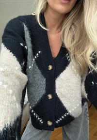 Fekete, szürke és krémszínű kötött kardigán gyémántmintákkal. Öt gombbal, széles ujjakkal és V-nyakú kialakítással rendelkezik. Puha tapintású.