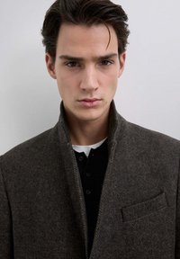 Abrigo de espiga marrón con un alto cuello, que presenta un bolsillo lateral. Debajo, una camisa negra con botones y una camiseta blanca visible.