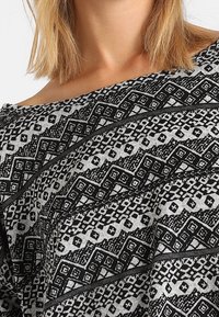 Zwart-wit gestippeld stof met geometrische ontwerpen. Zachte textuur, off-shoulder snit en contrasterende lijnen die een gelaagd effect creëren.