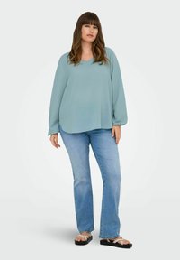 Svetlo modra dolgi rokav vrh z V-izrezom, v kombinaciji z razširjenimi modrimi džinsami. Vrh ima gladko teksturo; džins hlače imajo rahlo bledenje.