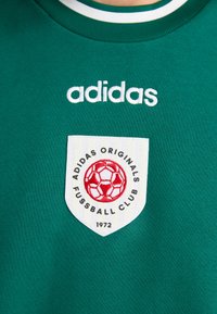 Grønn bomullssweatshirt med hvit ribbestrikket hals, brodert "adidas"-logo og merket med rød fotballdesign og klubbdetaljer.