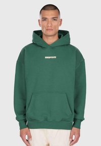 Un homme aux cheveux courts portant un sweat à capuche vert forêt avec le texte "DROPSIZE" sur la poitrine et un pantalon crème, se tenant devant un fond clair.
