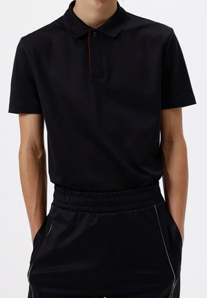 Black polo shirt with a collar and a small red accent on the placket, paired with black elasticated trousers featuring green side piping.