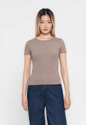 Femme jeune avec des cheveux droits à longueur d'épaules portant un haut à manches courtes en maille taupe et un jean bleu foncé, se tenant devant un fond blanc.