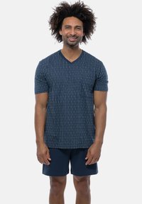 Marineblå V-hals t-shirt med lodrette hvide striber og en brystlomme, som er parret med marineblå shorts, lavet af blødt stof. Model står med afslappede arme.