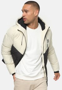Puffers jacket med en cremefarvet overdel og sort nederste del. Har hætte, lynlåslukning og tekstureret stof. Sorte detaljer på kraven.