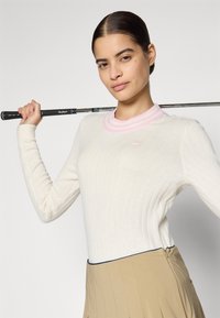 Pull crème à manches longues à côtes avec un col à rayures roses. Le mannequin tient un club de golf, portant une jupe plissée beige avec une fermeture éclair.