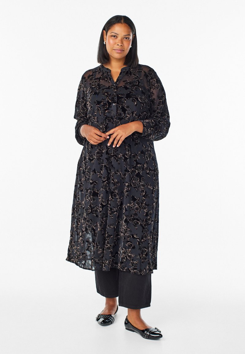 Črna prosojna obleka z dolgimi rokavi in cvetličnim čipkastim prekrivalom ter gumbastim izrezom. V kombinaciji z črnimi hlačami in sijočimi balerinskimi čevlji z dekorativnimi dodatki.