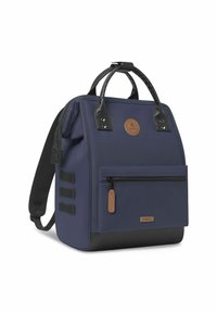 Zaino blu marino con manici in pelle nera, tasca frontale con zip e patch del logo marrone. Presenta un fondo nero testurizzato e un design minimalista.