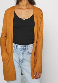 Dlouhý cardigan s otevřeným předním dílem v zlatavě oranžové barvě, pletená textura s vroubkovanými manžetami a bočními kapsami, nošený přes černé tílko a světlé džíny.