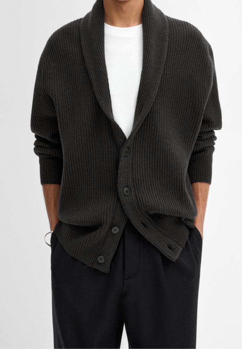 Cardigan côtelé foncé avec col châle, doté de six boutons, porté sur une chemise blanche, associé à un pantalon noir, mettant en valeur son design texturé.